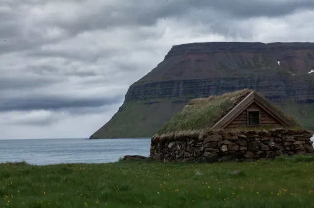 Hut with a turf roof and stone walls on the shore, with a steep-sided plateau across the water.