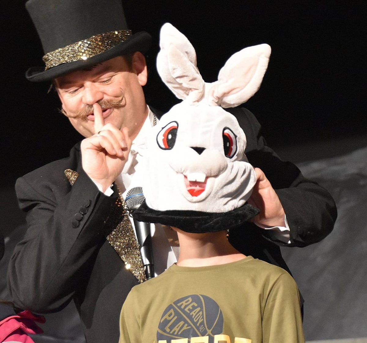 A performer in a top hat presents a large rabbit mask on stage.