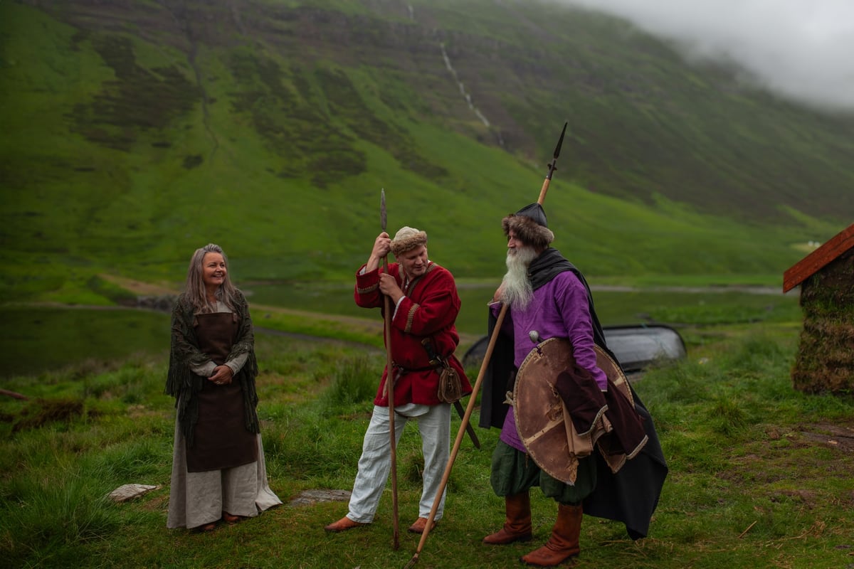 Three people in period dress on the grass in front of the skáli: a woman at the left, a man in a red tunic with a spear, a man in a purple cape with a shield and a spear