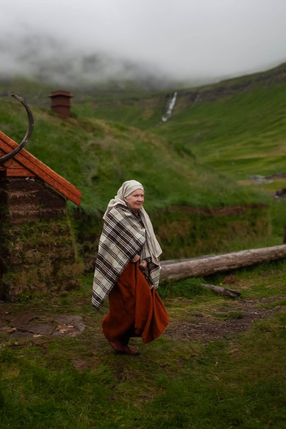 A woman in a headscarf and a rust-coloured skirt walks along a path beside the skáli; at the left the gable with a carved wooden ornament and a chimney