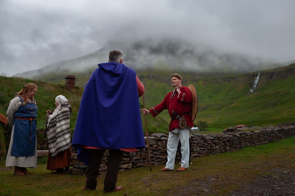 A group in period dress on the grass in front of the skáli; a man in a blue cloak stands with his back to the camera, facing a man in a red tunic holding a shield