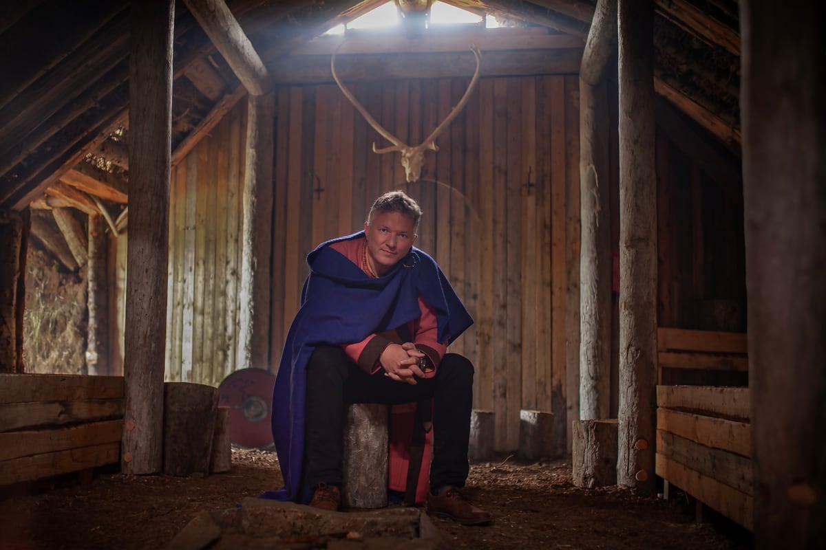 Eyþór Eðvarðsson sits inside the skáli, between the timber posts, wearing a blue woollen cloak; antlers hang on the wall behind him