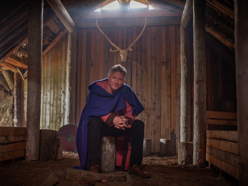 Eyþór Eðvarðsson sits inside the skáli, between the timber posts, wearing a blue woollen cloak; antlers hang on the wall behind him