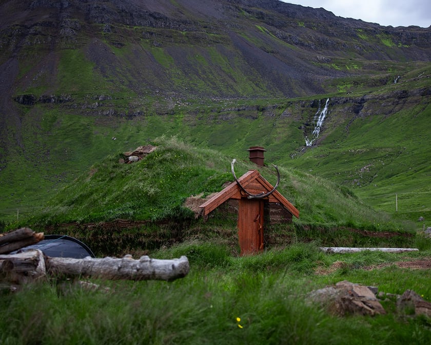 Skáli Hallvarðs Súganda seen from the front: a turf roof, a red gable with an iron ring, a boat and logs at the left