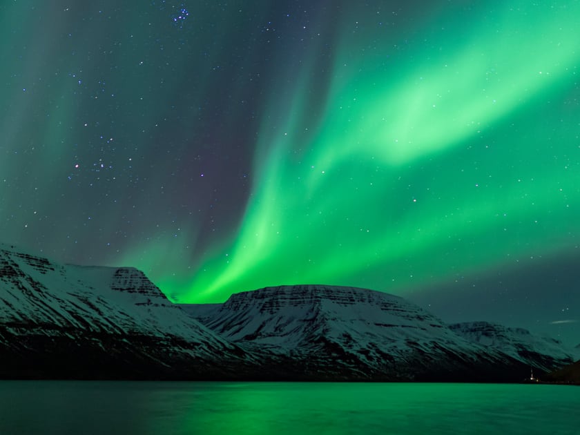 Northern lights over Súgandafjörður: green streaks across a starry sky above snow-covered mountains and the still water of the fjord.